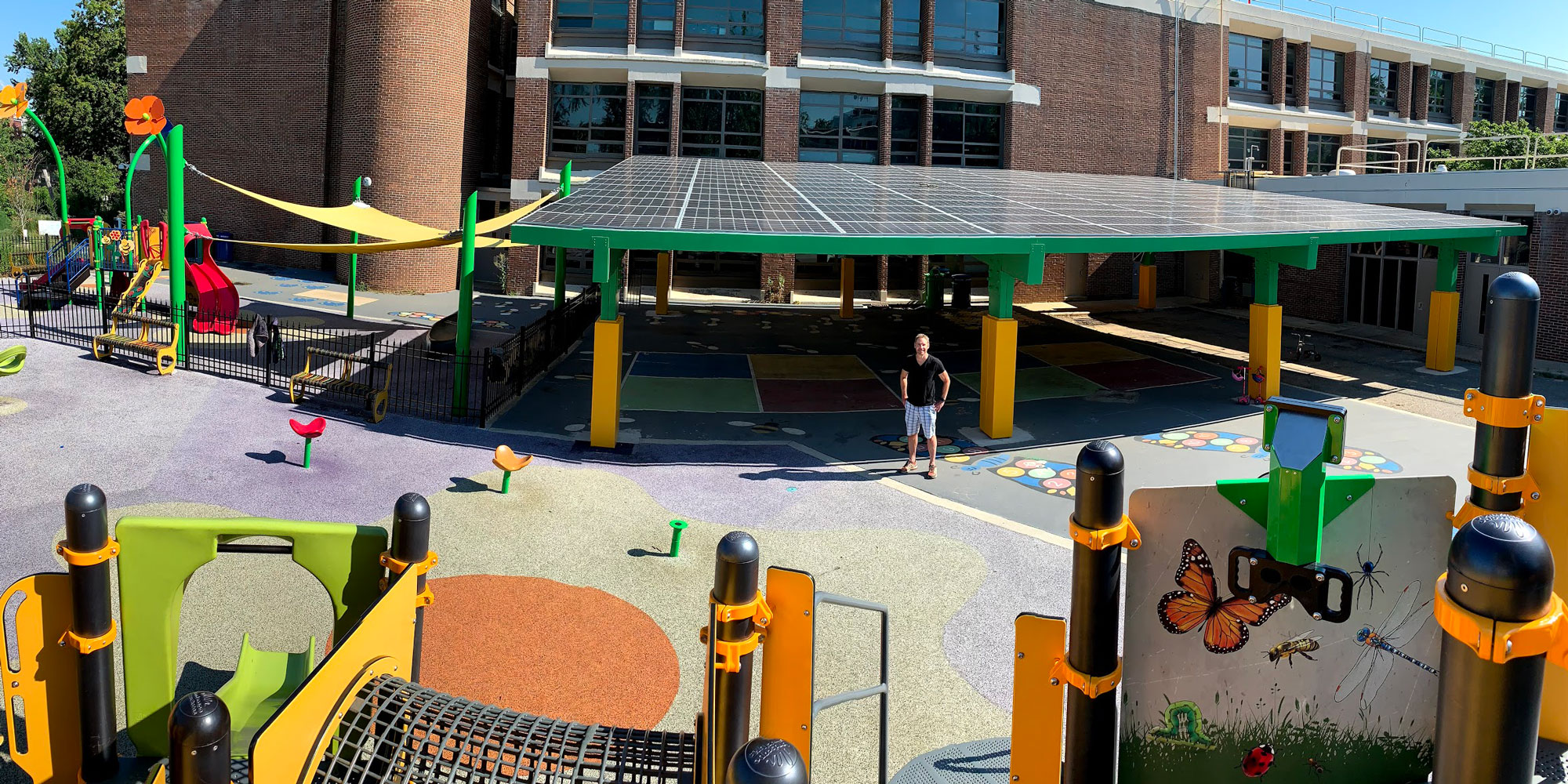 A person is standing outside of Ludlow-Taylor Elementary School. The school is a brick building with glass panes surrounded by trees and a bright, clear, sky. There is a bright playground structure with an illustration of insects on it. Left of the person is a series of flower-like playground structures staggered to look like a flower blooming. Beneath them is an illustration on the concrete resembling the sun with round edges instead of pointed rays. Behind the structures is a tarp covered area with a short metal fence. Behind the person and to the right of the tarp covered area are two four square areas which are covered by a large, bright solar panel area acting as a roof. To the right of the person are colorful illustrations on the ground designed to look like two curvy stone pathway.