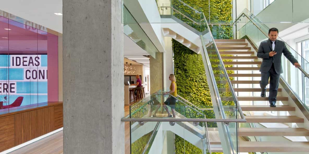 A person walking down smooth, back-less, stairs with glass panes and cylindrical stair rails on each side. In the backdrop is a vertical wall of greenery and two other people are in the backdrop of the office building. There is a concrete pillar that the stairs are connected to and to the left is what appears to be a conference room with slightly tinted windows and a background that says 'Ideas'