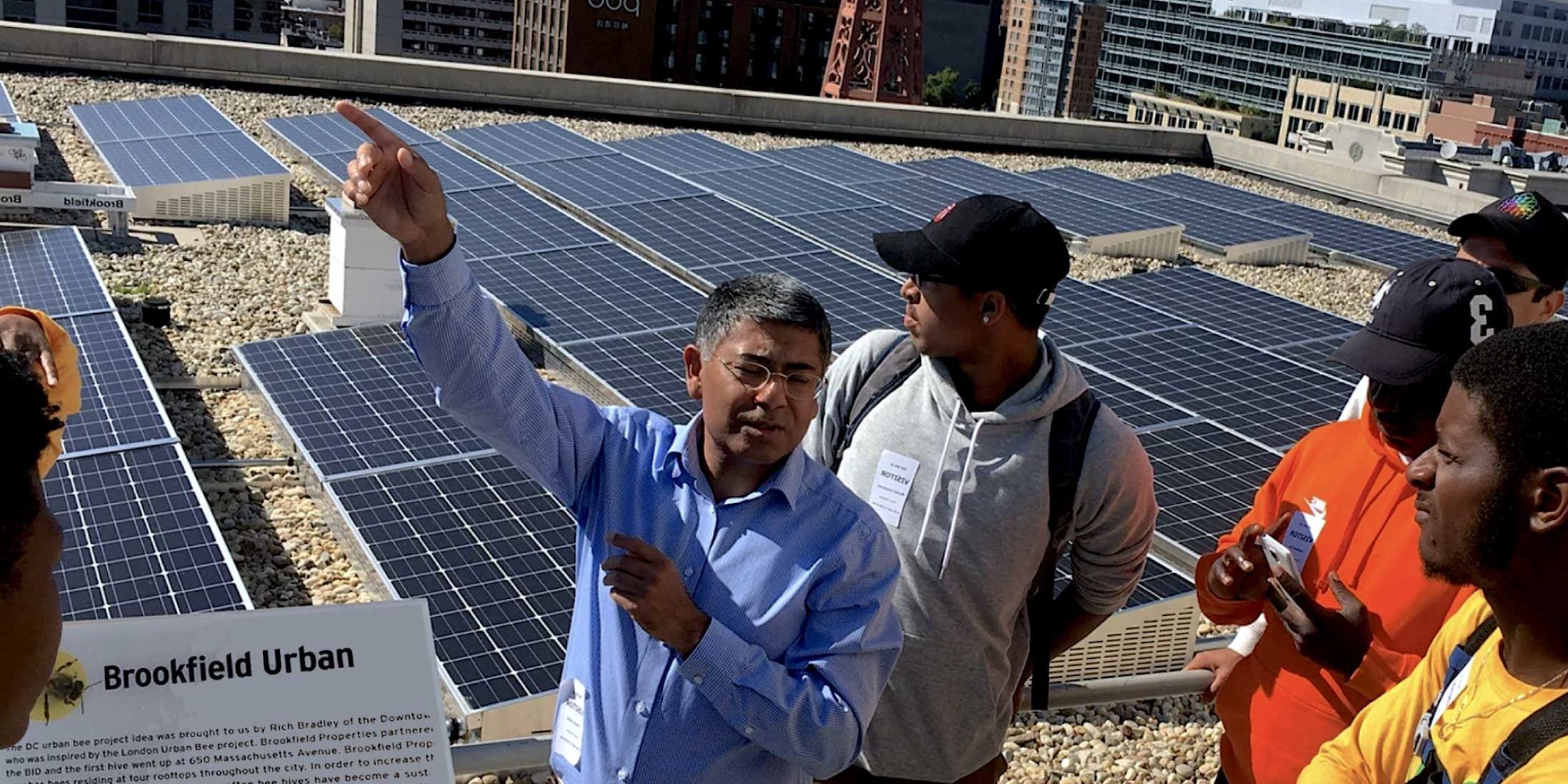 A group of about five people with one person on the side standing in front of solar panels on an outdoor rooftop. The rooftop overlooks a city in the backdrop. A sign that says 'Brookfield Urban' with further text below is to the left of the people.