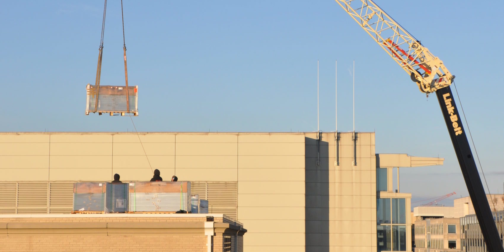 Three people are standing behind panels packages of panels on top of a building. A crate suspended by a crane is above them. Behind them is a building and to the right is the crane. Buildings are in the backdrop on the right and there is a clear, bright sky.