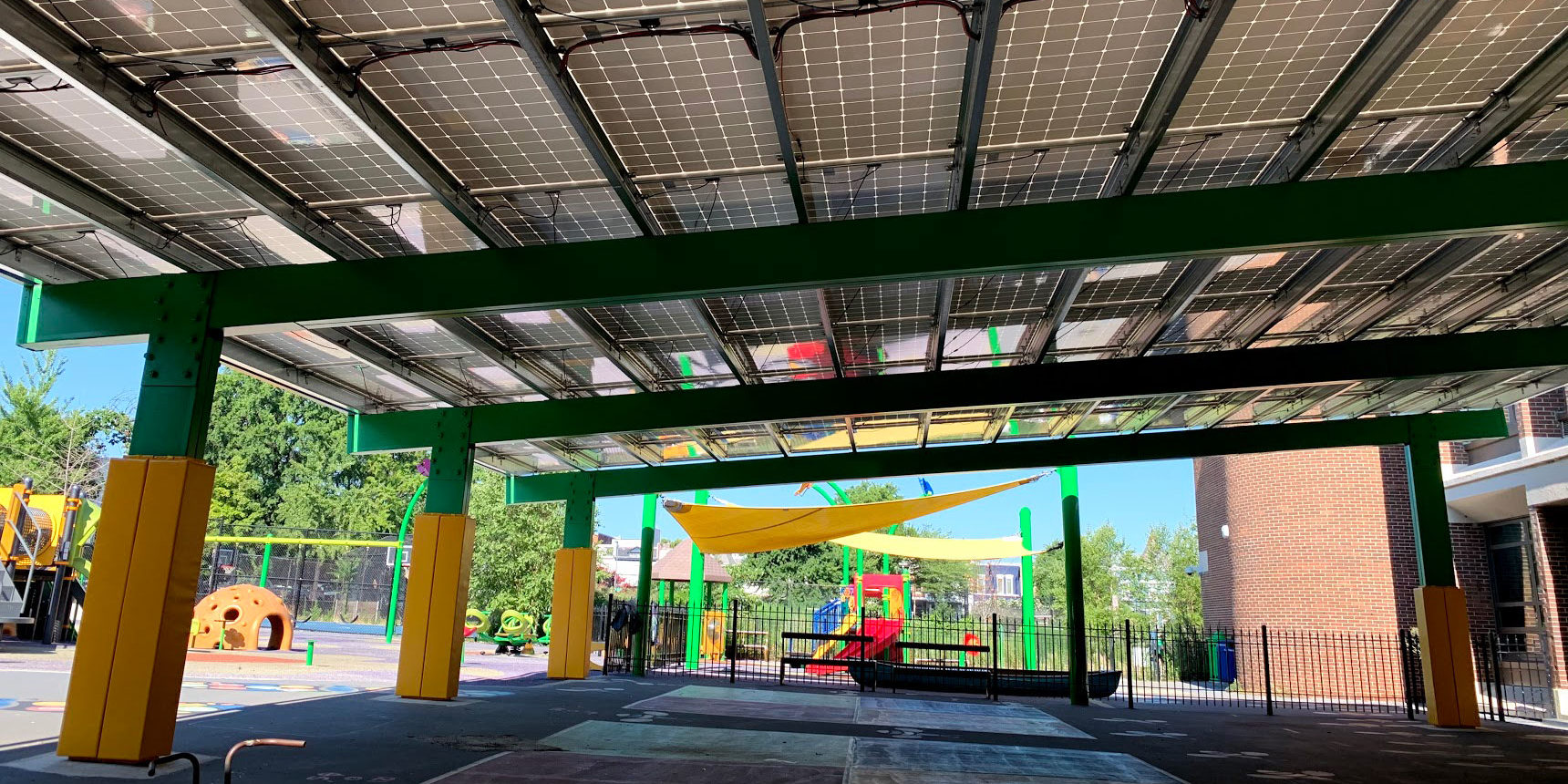 An eye-level view of Ludlow-Taylor Elementary School's solar panel roof. The photo appears to be taken under the roof and the four-square chalk area on the concrete is in front. Supportive pillars are on the left as well as a playground. Another playground set is in the backdrop behind a short iron fence. To the right is a brick building and further behind are trees, buildings, and a clear, sunny sky.