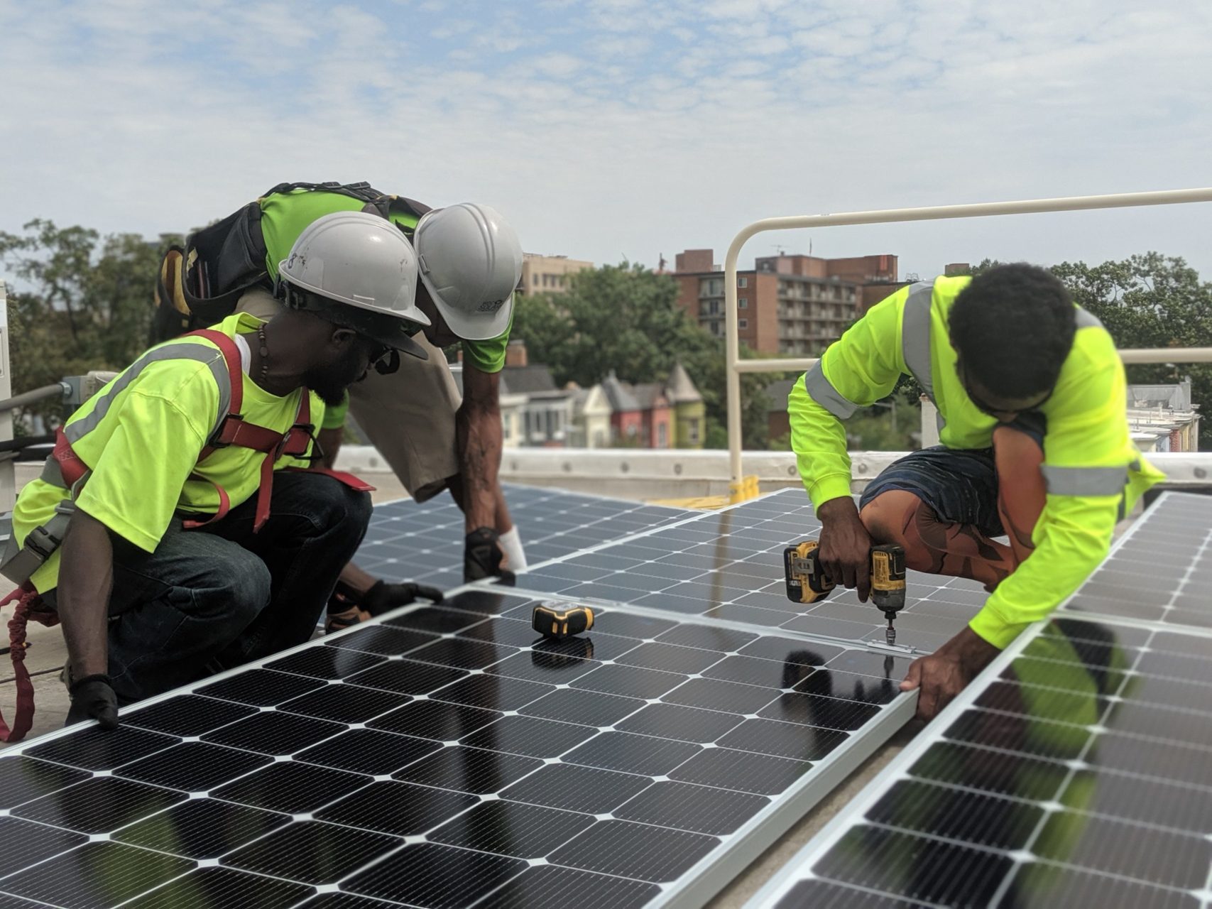 Three people standing on a rooftop around solar panels. Two are wearing construction hats to the left and the person on the right is holding a drill. They are all wearing neon safety shirts. The backdrop has trees, houses, buildings, and the sky is partially cloudy but parts of it are bright.
