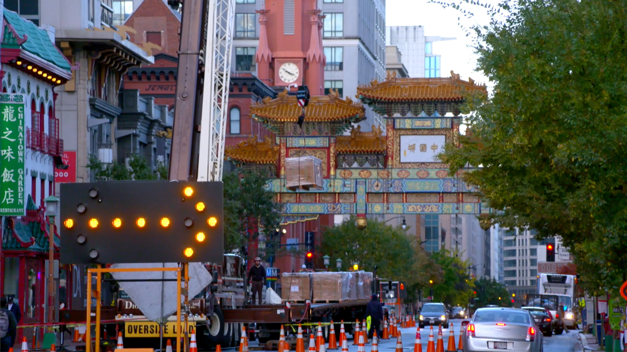 A neon construction arrow sign points right in front of a crane. The sign and crane are outside in front of a Chinatown community gate with many orange cones surrounding the crane. A sign to the left says 'Chinatown Garden Restaurant' and beyond the gate is a clock tower with four conical small towers surrounding the actual clock, and glass-paned buildings are behind the Chinatown community gate. There are cars on the road and trees lining the streets. The sky is bright with no outlines of clouds.